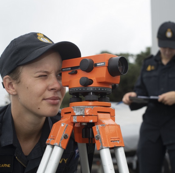 A Royal New Zealand Navy hydrographers being assessed by another Navy sailor in the background. The sailor is looking through a piece of equipment that is orange, they have one eye looking through the scope and the other closed.  