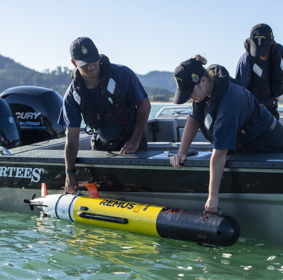 Three Royal New Zealand Navy personnel in a boat on a nice day. Two sailors lean over the boat to reach for a piece of equipment. In the background there are tree covered hills. 
