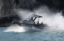 Royal New Zealand Navy Pathfinder boat moves through the water at speed on a sunny day. In the background you see the edge of a cliff