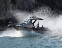 Royal New Zealand Navy Pathfinder boat moves through the water at speed on a sunny day. In the background you see the edge of a cliff