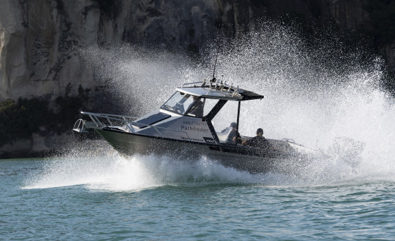 Royal New Zealand Navy Pathfinder boat moves through the water at speed on a sunny day. In the background you see the edge of a cliff
