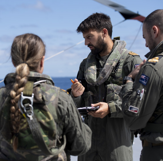 Four Royal New Zealand Navy aircraft personnel talking in a group. The person whose face is shown is holding a pen and a notepad. 