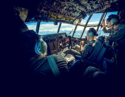 The Royal New Zealand Air Force aircrew on board one of the C-130H(NZ) Hercules aircraft during a flight. Outside the window of the aircraft you can see land and a sunny day with some cloud. Within the flight deck there is a pilot close to the left side o