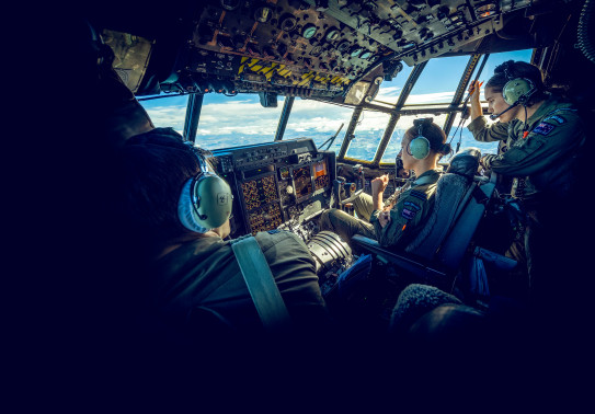 The Royal New Zealand Air Force aircrew on board one of the C-130H(NZ) Hercules aircraft during a flight. Outside the window of the aircraft you can see land and a sunny day with some cloud. Within the flight deck there is a pilot close to the left side o