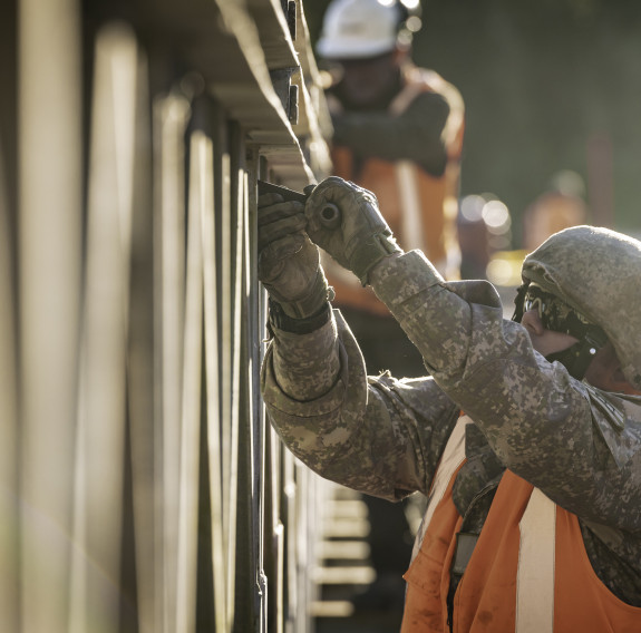 A New Zealand Army soldier works on a bridge with a helmet, glasses and an orange high vis vest. There are other people in the background working on the bridge as well.