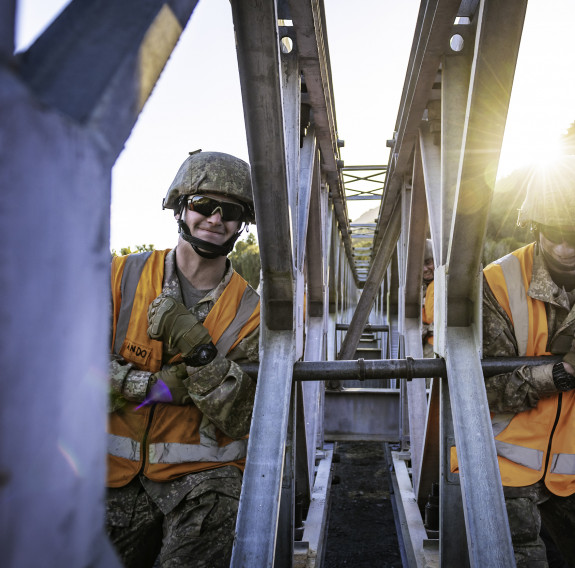 Two New Zealand Army soldiers lift part of a Bailey Bridge, one on each side of it. The image is backlit with sun and has been taken in the centre of the bridge.