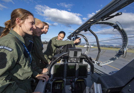 Three School to Skies candidates look into the cockpit of one of the T-6C Texan II as a pilot explains the features. It's a nice day with some clouds and blue skies. 