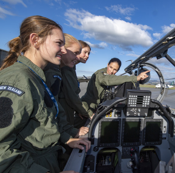 Three School to Skies candidates look into the cockpit of one of the T-6C Texan II as a pilot explains the features. It's a nice day with some clouds and blue skies. 