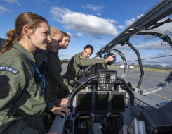 Three School to Skies candidates look into the cockpit of one of the T-6C Texan II as a pilot explains the features. It's a nice day with some clouds and blue skies. 