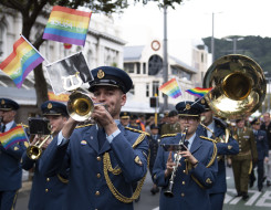 Royal New Zealand Air Force Band members walk down a street playing their instruments with rainbow flags. 