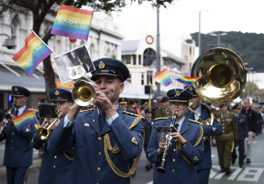 Royal New Zealand Air Force Band members walk down a street playing their instruments with rainbow flags. 