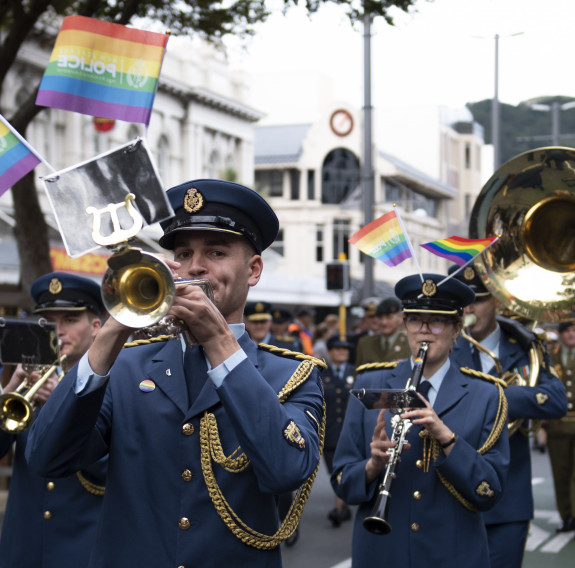 Royal New Zealand Air Force Band members walk down a street playing their instruments with rainbow flags. 