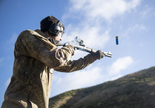 A soldier fires a Benelli M3 (NZ) Tactical Shotgun. The photo is shot from a low angle looking up to the soldier from the side. The soldier is wearing hearing protection and you can see the bullet coming out. There is a hill in the background. 