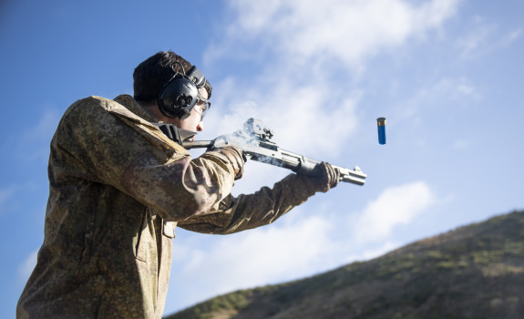 A soldier fires a Benelli M3 (NZ) Tactical Shotgun. The photo is shot from a low angle looking up to the soldier from the side. The soldier is wearing hearing protection and you can see the bullet coming out. There is a hill in the background. 