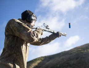 A soldier fires a Benelli M3 (NZ) Tactical Shotgun. The photo is shot from a low angle looking up to the soldier from the side. The soldier is wearing hearing protection and you can see the bullet coming out. There is a hill in the background. 