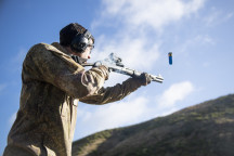 A soldier fires a Benelli M3 (NZ) Tactical Shotgun. The photo is shot from a low angle looking up to the soldier from the side. The soldier is wearing hearing protection and you can see the bullet coming out. There is a hill in the background. 