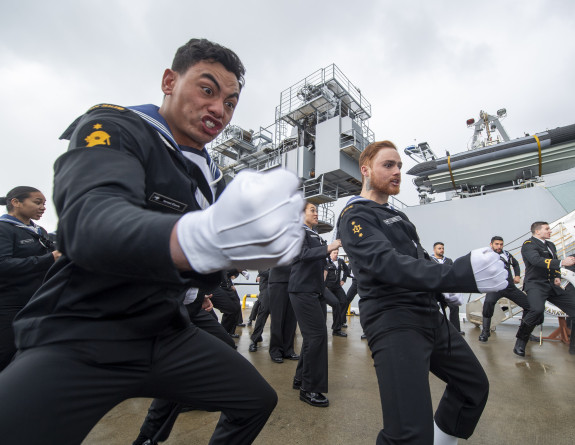Royal New Zealand Navy sailors perform a haka near the ship. The sailor in the foreground in focus has a fist and some strong enthusiastic emotion on their face.