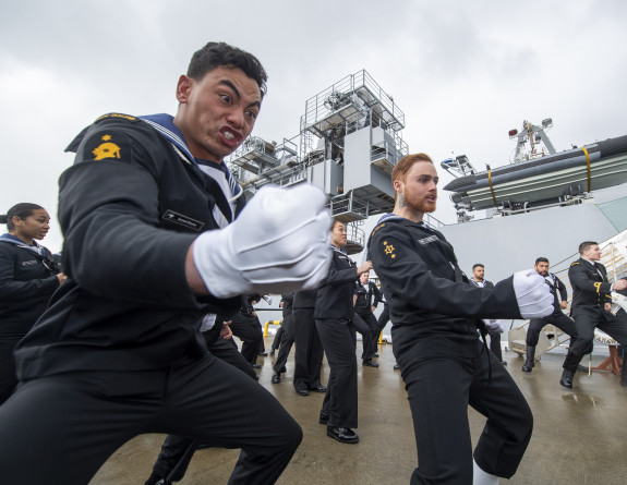 Royal New Zealand Navy sailors perform a haka near the ship. The sailor in the foreground in focus has a fist and some strong enthusiastic emotion on their face. 