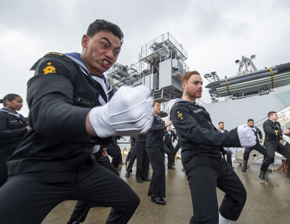 Royal New Zealand Navy sailors perform a haka near the ship. The sailor in the foreground in focus has a fist and some strong enthusiastic emotion on their face.