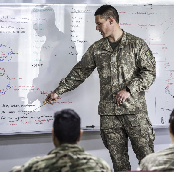 A New Zealand Army soldier points a whiteboard with content on it. In the foreground, blurred are other uniformed people watching on. 