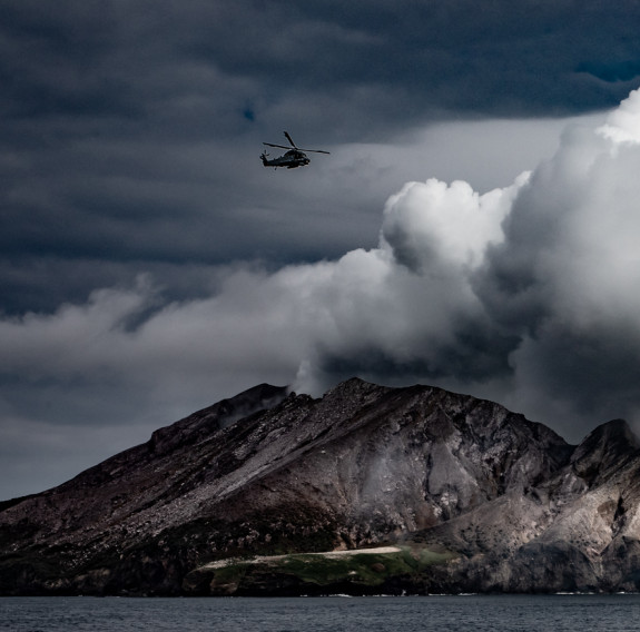 A SH-2G(I) Seasprite Helicopter flying above a thick plume of white smoke coming from Whaakari/White Island following the eruption.