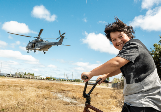In the foreground a young child smiles while sitting on his bike out of excitement seeing the Royal New Zealand Air Force NH90 helicopter flying in the background. It's a sunny summer day with some cloud and dry grass.