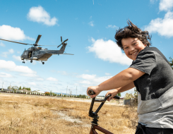 In the foreground a young child smiles while sitting on his bike out of excitement seeing the Royal New Zealand Air Force NH90 helicopter flying in the background. It's a sunny summer day with some cloud and dry grass.