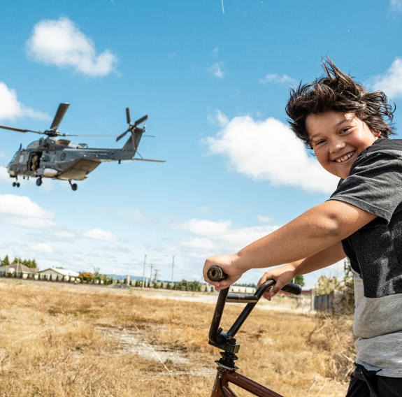 In the foreground a young child smiles while sitting on his bike out of excitement seeing the Royal New Zealand Air Force NH90 helicopter flying in the background. It's a sunny summer day with some cloud and dry grass.