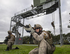 New Zealand Army soldiers fast roping from a tower in the background and in the foreground two soldiers provide ground cover below the tower. One solider on the tower watches as and accesses the soldiers..
