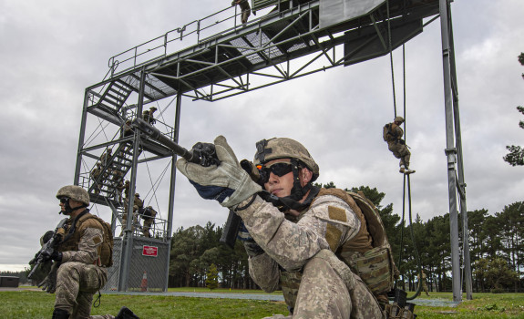 New Zealand Army soldiers fast roping from a tower in the background and in the foreground two soldiers provide ground cover below the tower. One solider on the tower watches as and accesses the soldiers..