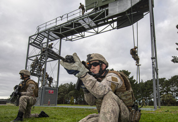 New Zealand Army soldiers fast roping from a tower in the background and in the foreground two soldiers provide ground cover below the tower. One solider on the tower watches as and accesses the soldiers..