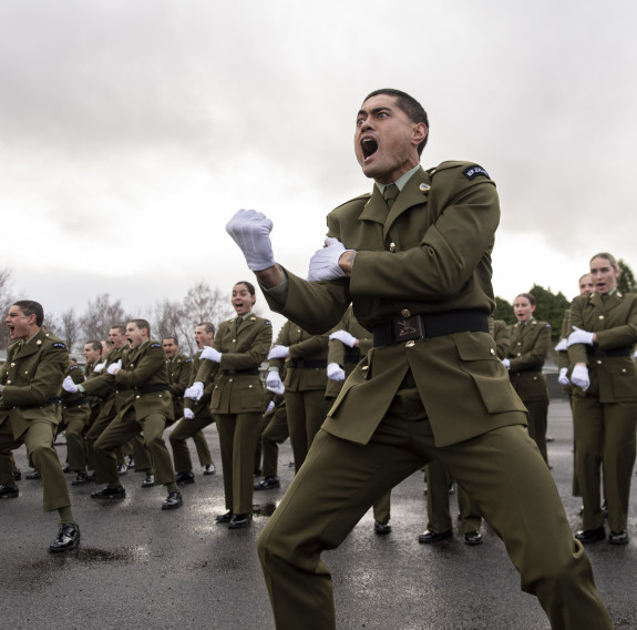 New Zealand Army soldiers perform a haka. The soldier in the foreground is making a fist as part of the haka and the photo is caught while yelling. Other soldiers are seen in the background.