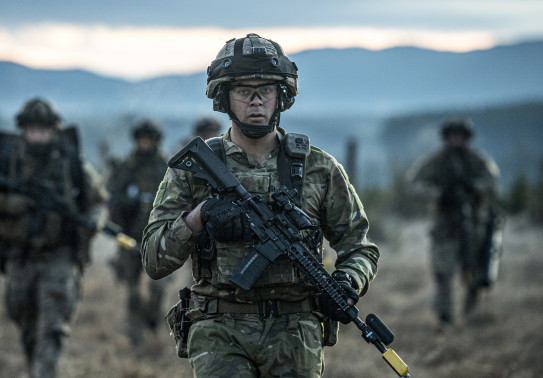 A New Zealand Army soldier in uniform in the foreground carries a MARS-L weapon with fellow soldiers in the background