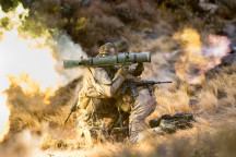 New Zealand Army soldiers fire a Carl Gustaf M3 there are flames out the back and motion shown out the front of the weapon. the soldiers are training in the tussock grass
