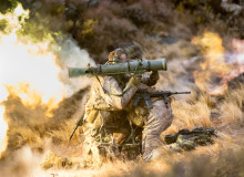 New Zealand Army soldiers fire a Carl Gustaf M3 there are flames out the back and motion shown out the front of the weapon. the soldiers are training in the tussock grass