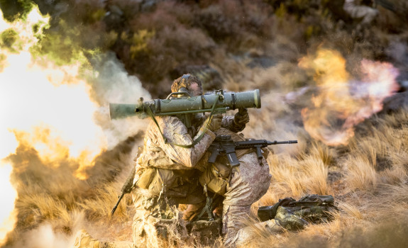 New Zealand Army soldiers fire a Carl Gustaf M3 there are flames out the back and motion shown out the front of the weapon. the soldiers are training in the tussock grass