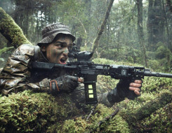 A New Zealand Army soldier in the New Zealand bush with MARS-L weapon. The area is all wet and very green. The soldier has his mouth open as he yells a command. 