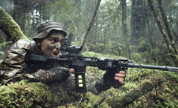 A New Zealand Army soldier in the New Zealand bush with MARS-L weapon. The area is all wet and very green. The soldier has his mouth open as he yells a command. 