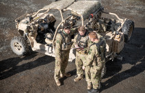 An aerial photo of three soldiers standing next to a Polaris MRZR vehicle. The three soldiers are discussing something and one soldier is pointing to a notepad