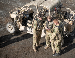 An aerial photo of three soldiers standing next to a Polaris MRZR vehicle. The three soldiers are discussing something and one soldier is pointing to a notepad