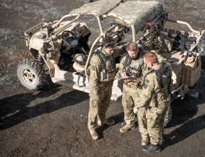 An aerial photo of three soldiers standing next to a Polaris MRZR vehicle. The three soldiers are discussing something and one soldier is pointing to a notepad