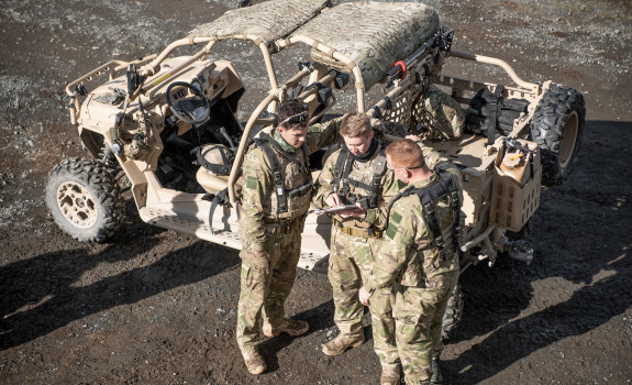 An aerial photo of three soldiers standing next to a Polaris MRZR vehicle. The three soldiers are discussing something and one soldier is pointing to a notepad