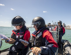 School to Seas candidates on the front of a Royal New Zealand Navy Rigid Hulled Inflatable Boat (RHIB) in Auckland Harbour, sailors in the background driving the boat and Auckland city in the distant background. It's a sunny day with blue sky. 
