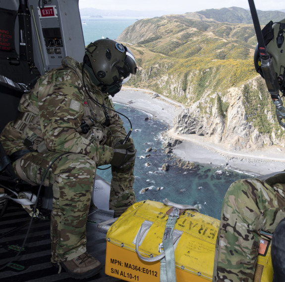 Royal New Zealand Air Force helicopter loadmasters look outside the aircraft during a search and rescue mission. In the photo you can see the rigid coastline and ocean. There is also a box at their feet they would use if they found the missing people