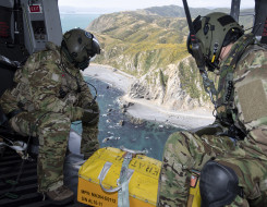 Royal New Zealand Air Force helicopter loadmasters look outside the aircraft during a search and rescue mission. In the photo you can see the rigid coastline and ocean. There is also a box at their feet they would use if they found the missing people