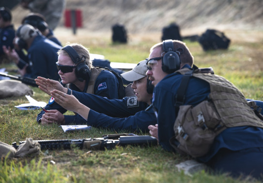 Royal New Zealand Air Force personnel work together on the rifle range. One of the personnel signals in the direction to the left while the other two look in that direction. All personnel are wearing hearing protection.