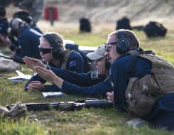 Royal New Zealand Air Force personnel work together on the rifle range. One of the personnel signals in the direction to the left while the other two look in that direction. All personnel are wearing hearing protection.