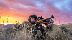 Two New Zealand Army soldiers lie in the tussock grass with the MAG 58 - 7.62mm Machine Gun. One soldier looks through the scope and the other looks toward the target. The sky is pink and yellow sunset skies. 