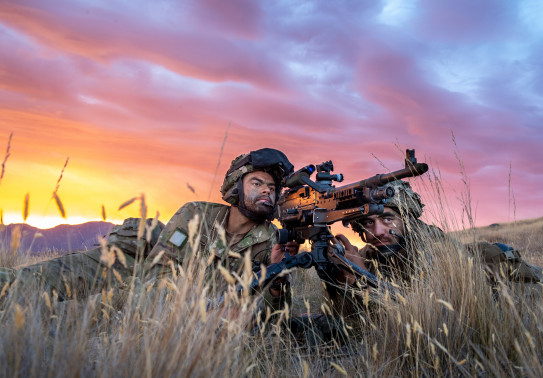 Two New Zealand Army soldiers lie in the tussock grass with the MAG 58 - 7.62mm Machine Gun. One soldier looks through the scope and the other looks toward the target. The sky is pink and yellow sunset skies. 