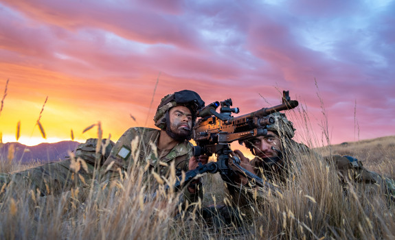 Two New Zealand Army soldiers lie in the tussock grass with the MAG 58 - 7.62mm Machine Gun. One soldier looks through the scope and the other looks toward the target. The sky is pink and yellow sunset skies. 
